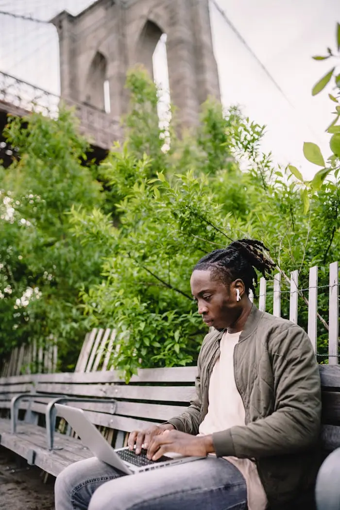 Young man focused on laptop work sitting on a park bench under a bridge.
